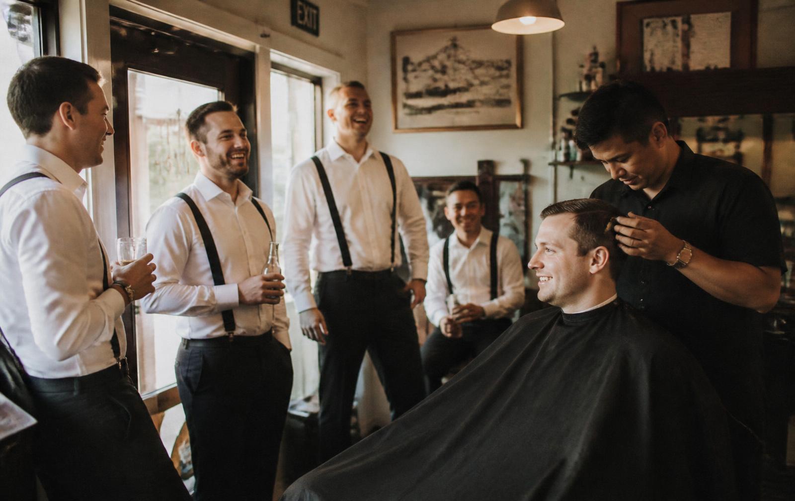 Groomsmen laughing together in the barbershop while the groom gets his haircut